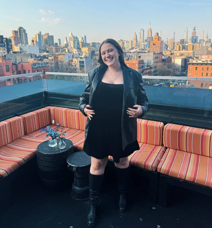 A woman stands smiling on a rooftop with a city skyline in the background. She is wearing a black outfit with a leather jacket and boots