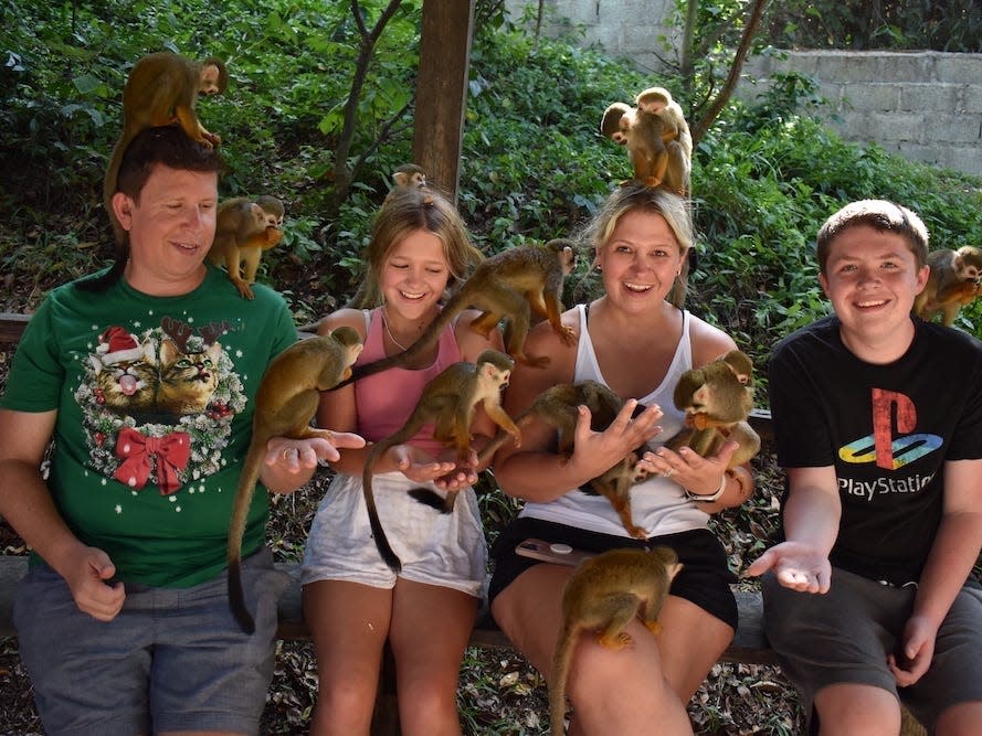 Terri Peters and her family sit on a wooden fence, with monkeys sitting on their shoulders and heads. Terri's husband wears a green shirt with cats wearing a Santa hat and faux antlers and blue shorts. Terri's daughter wears a pink tank top and white shorts. Terri wears a white tank top and black shorts. Terri's son wears a black t-shirt with the "Playstation" logo and grey shorts.