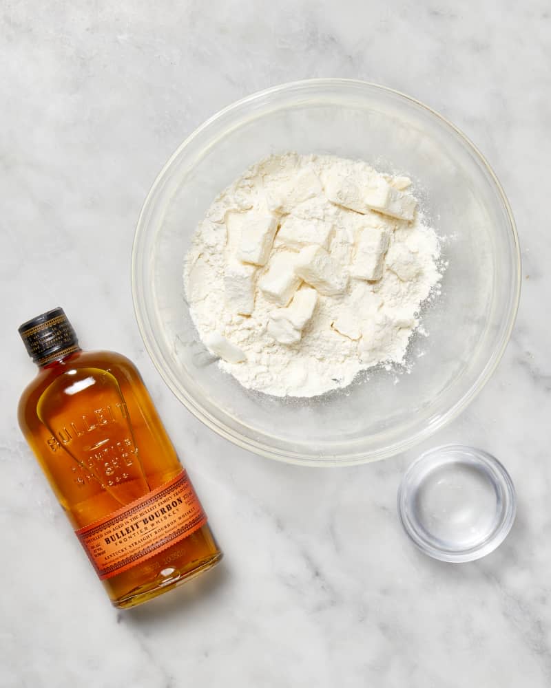 A bowl with flour and cubes of butter and a bottle of bourbon to make a pie crust seen from above.