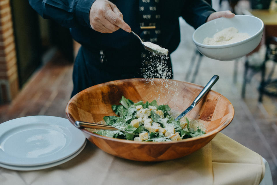 A person in dark clothing prepares a salad by sprinkling cheese over a wooden bowl filled with lettuce and croutons