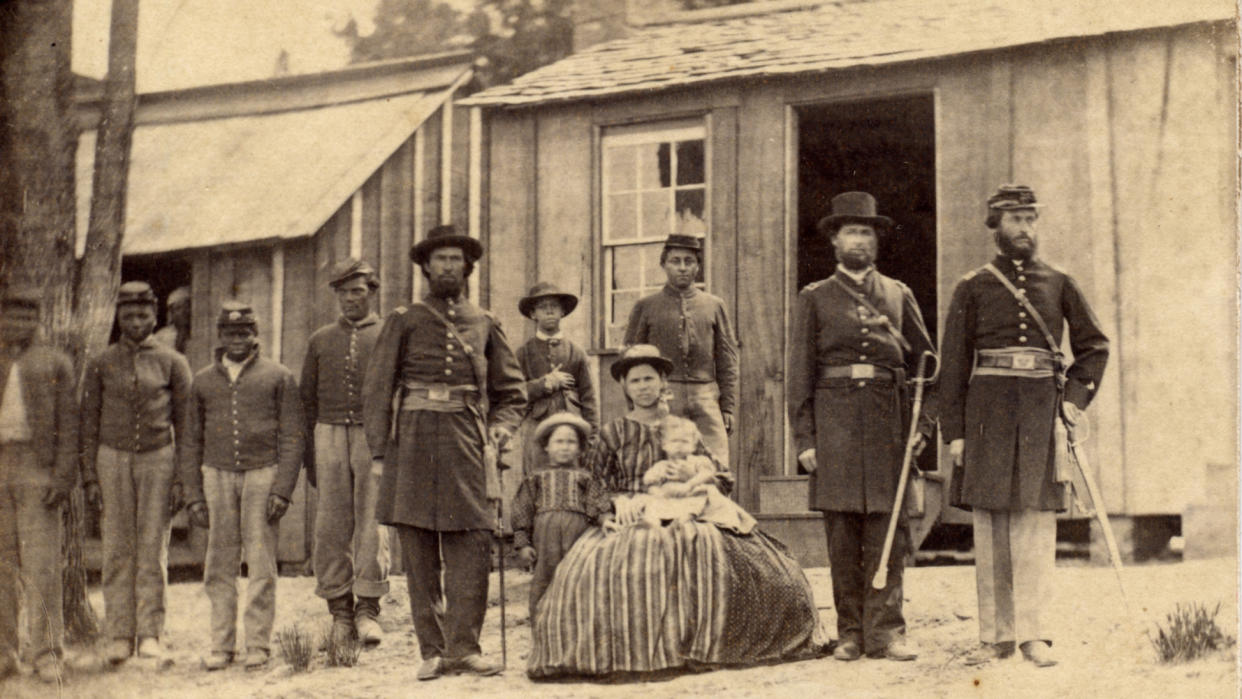 A dozen people pose in a row in front of a wooden building, including six African Americans, five in Union uniforms, a seated woman in a voluminous skirt with a baby on her knee and a small boy at her side, and three Union officers in long coats, one in a top hat, one in a Union cap, and one in a fedora.
