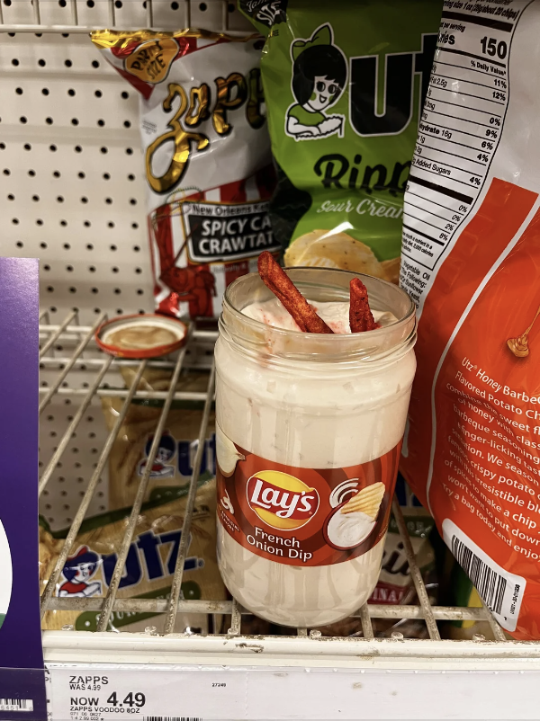 Jar of Lay's French Onion Dip with two snack sticks in it, on a grocery store shelf with various snack bags around