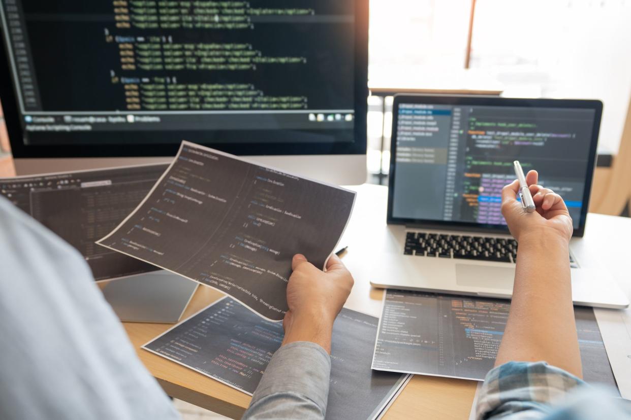 Focus on hands of two male software developers working at a desk with a desktop computer and a laptop with sheets of code and code on the screens with window and office in the background