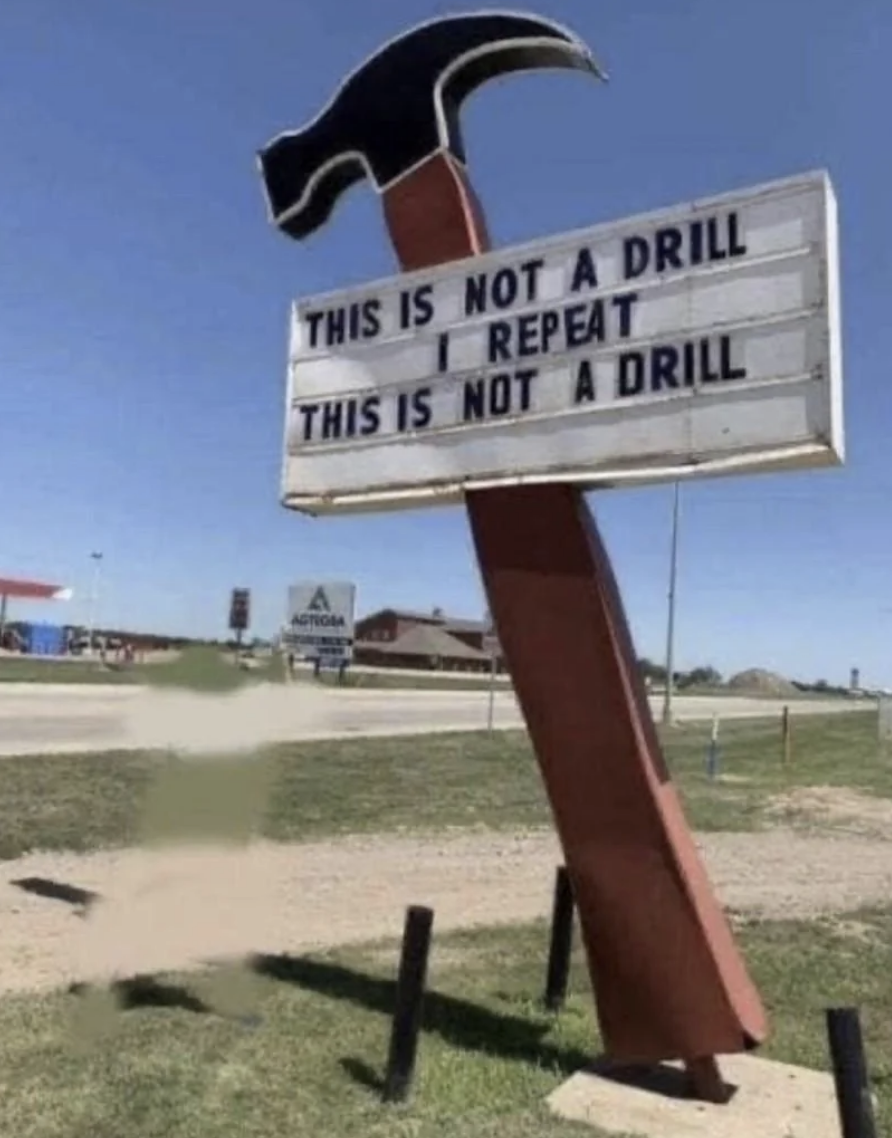 Sign shaped like an upside-down hammer with text "THIS IS NOT A DRILL" placed above a spinning drill bit creating a hole