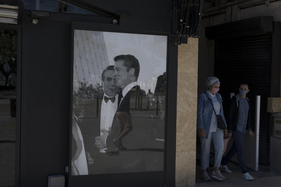 A couple wearing masks to protect from coronavirus walk by a poster of Brad Pitt and George Clooney in Cannes, southern France, Tuesday, May 12, 2020. The Cannes Film Festival won't kick off as planned on Tuesday. The festival's 73rd edition has been postponed indefinitely, part of the worldwide shutdowns meant to stop the spread of the coronavirus. (AP Photo/Daniel Cole)