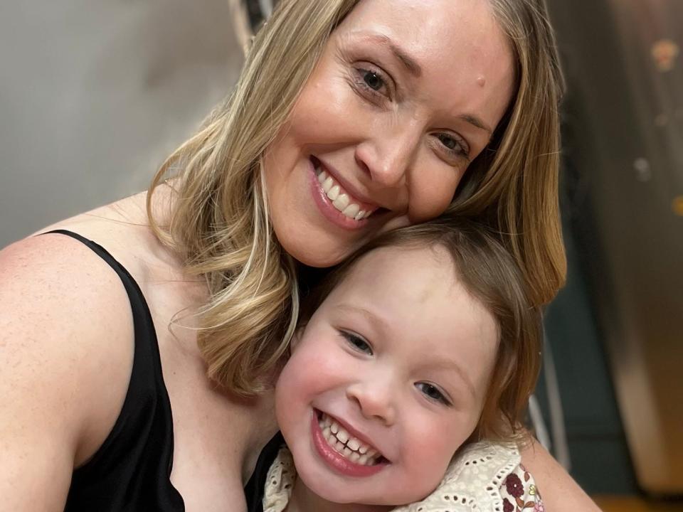 A woman hugging her young daughter with some balloons behind.