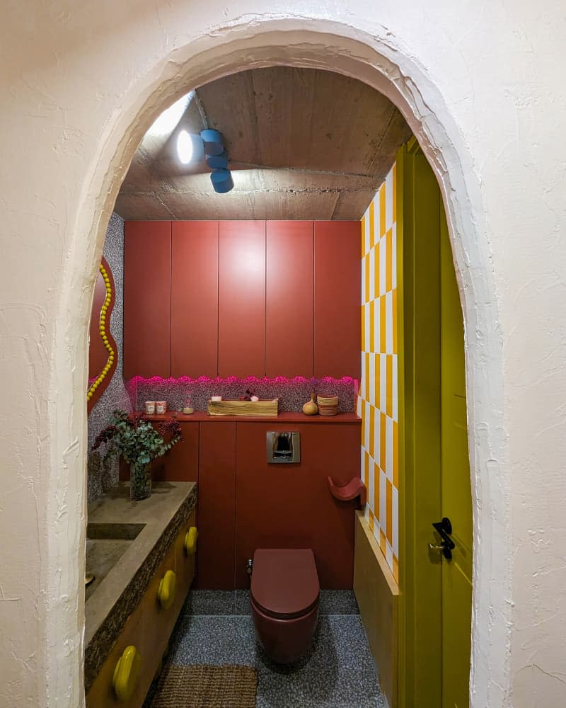 Bathroom seen through archway with wavy orange and yellow mirror, wood cabinets with stone slab counter and yellow irregular shaped knobs, and lots of orange cabinets