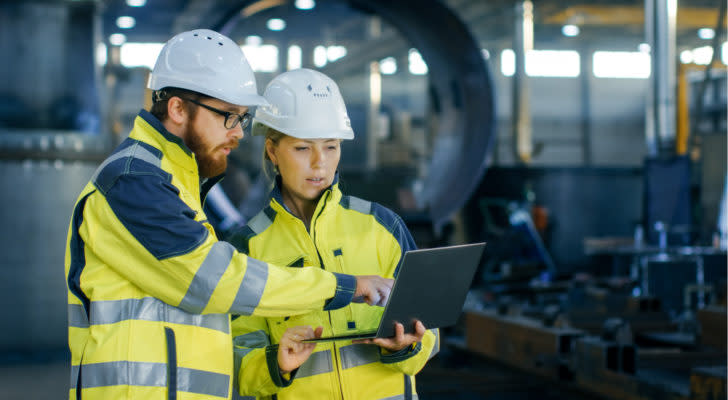 Engineers in yellow jackets and hard hats looking at a laptop in a workshop