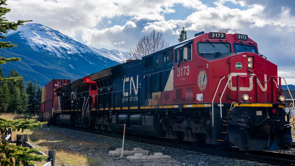 A freight train zips through a field. Snow-capped mountains are behind the train.