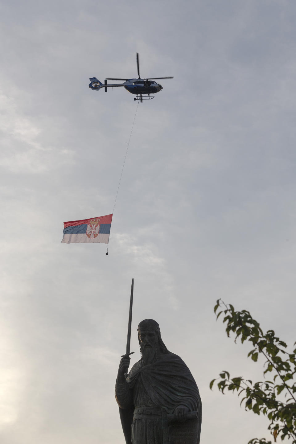 A helicopter carries the flag of Serbia as it passes the collosal monument to Stefan Nemanja, a Serbian medieval ruler, during a ceremony to mark the newly established "Day of Serb Unity, Freedom and the National Flag" state holiday in Belgrade, Serbia, Wednesday, Sept. 13, 2021. Serbia has kicked off a new holiday celebrating national unity with a display of military power, triggering unease among its neighbors. (AP Photo/Marko Drobnjakovic)