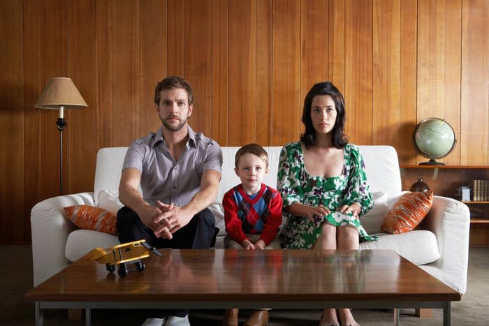 A family of three sits on a couch: man in a grey shirt, toddler in a plaid outfit, and woman in a green floral dress