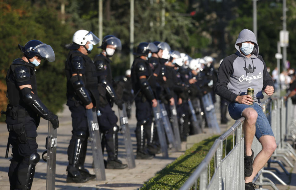 A man sits on a fence as the riot police guards the area in front of the Serbian Parliament building during a demonstration in Belgrade, Serbia, Wednesday, July 8, 2020. Serbia's president Aleksandar Vucic backtracked Wednesday on his plans to reinstate a coronavirus lockdown in Belgrade after thousands protested the move and violently clashed with the police in the capital. (AP Photo/Darko Vojinovic)