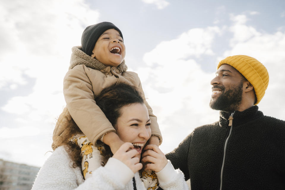 Child on woman's shoulders, man beside them smiling, outdoors, casual attire, family moment