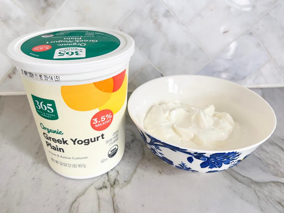 A light-yellow container of 365 organic plain Greek yogurt with a red, orange, and yellow design and green lid. The container sits next to a blue-and-white bowl filled with white yogurt