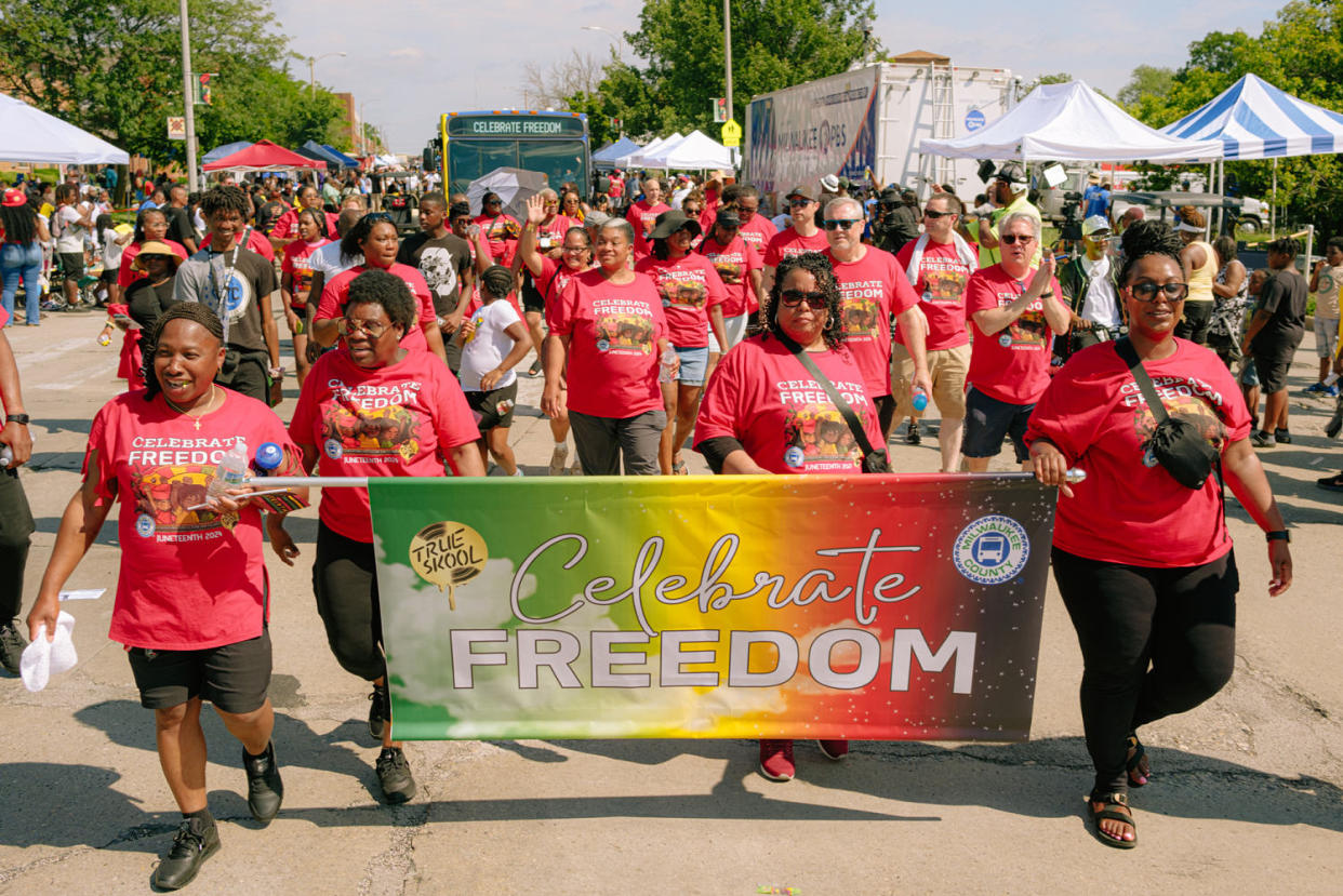 Parade goers walk on the street, holding a banner that says 