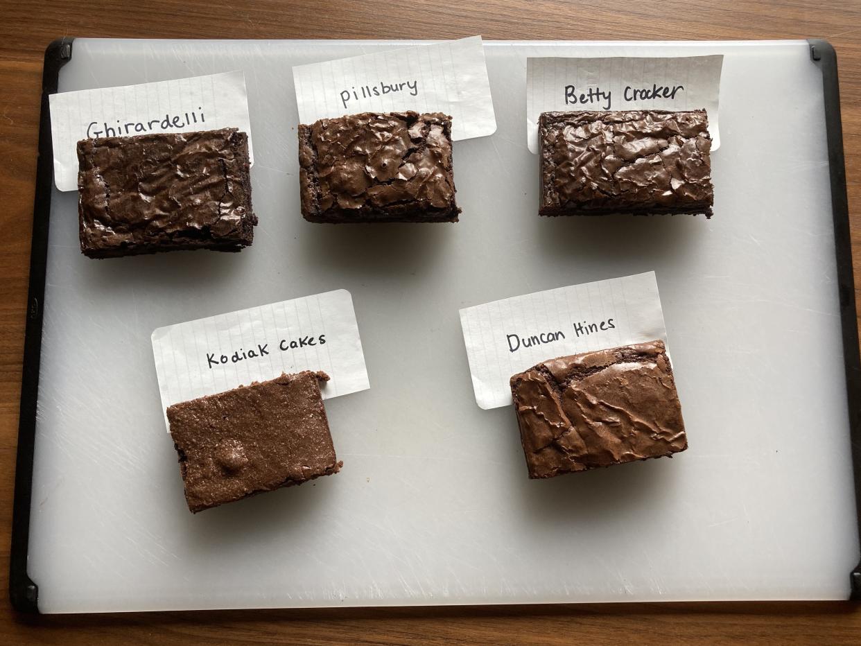 Five different brownie squares on a white cutting board with pieces of paper labeled "Ghirardelli," "Pillsbury," "Betty Crocker," "Kodak Cakes," and "Duncan Hines"