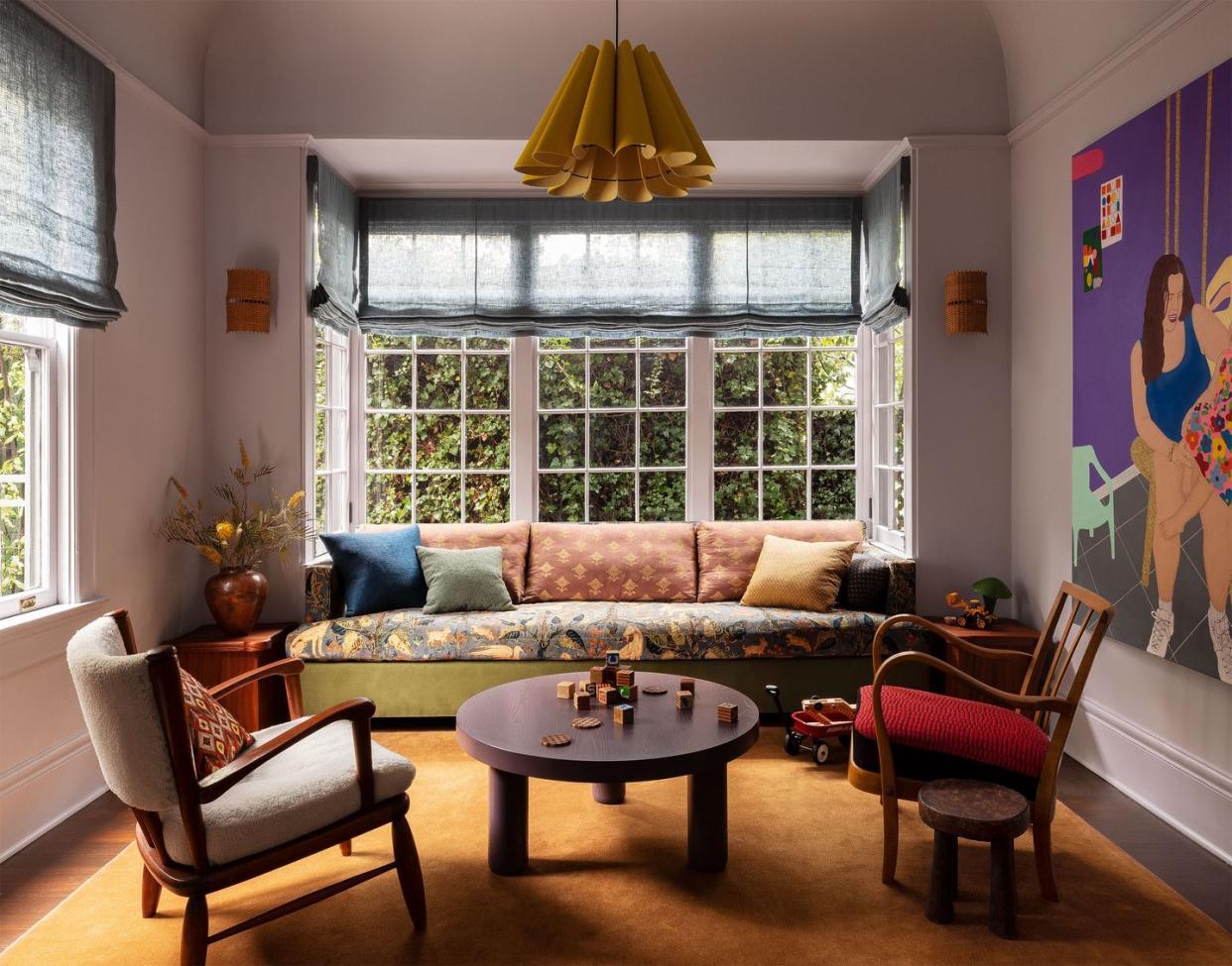 sofa with flowered seat and print back cushions and pillows set into bay window with roman shades, round cocktail table, wood chairs with cushion seats, fluted pendant, footstool, ocher rug, large artwork