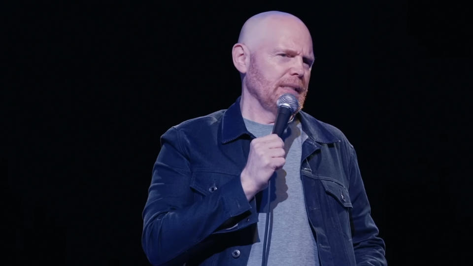 Bill Burr in a grey t-shirt and dark shirt over it, holding a microphone
