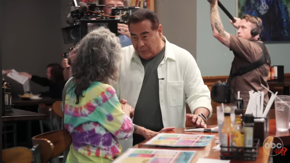 Two individuals at a diner table engaged in conversation; camera crew in background capturing the interaction