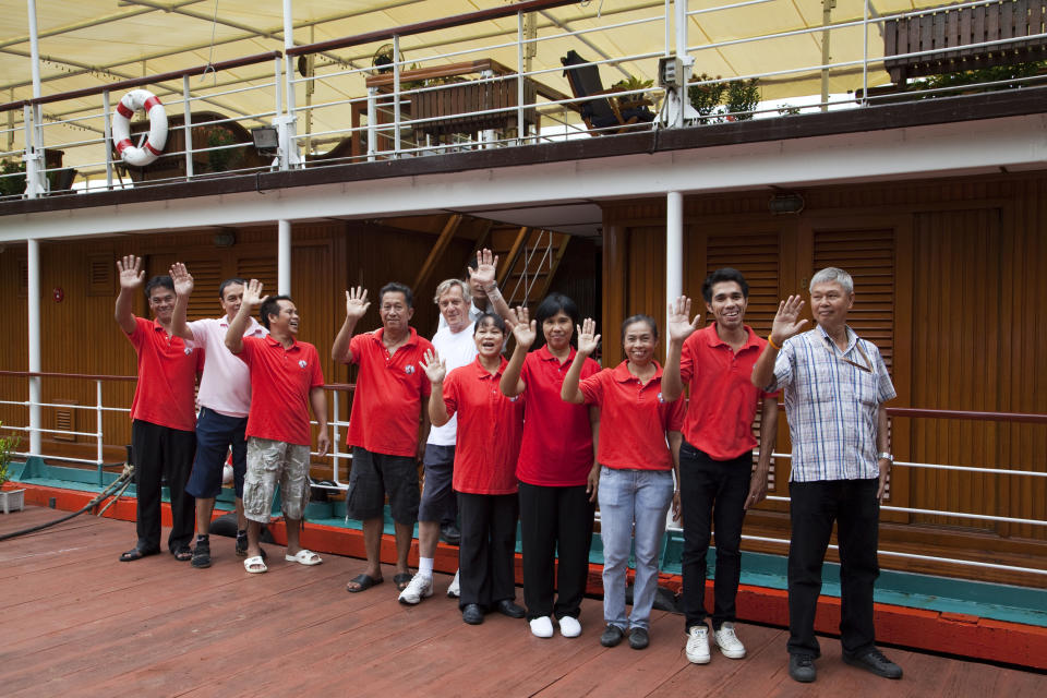 Group of nine people on a boat wearing casual red shirts, standing in a row, waving and smiling at the camera; one person on the right in a plaid shirt