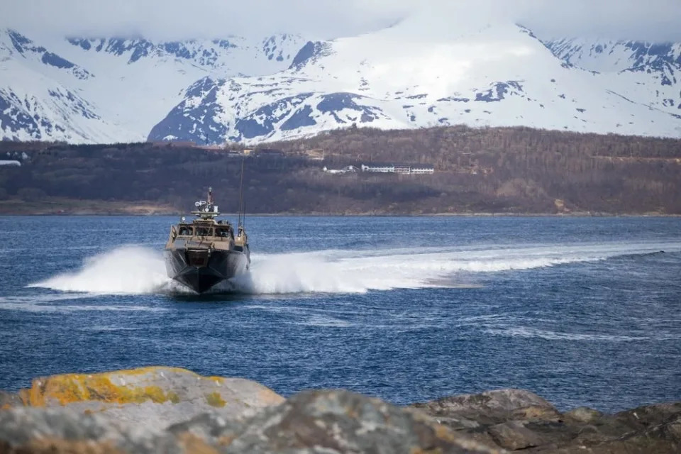 A CB90 fast assault boat approaches a rocky shore, with snowy mountains in the background.