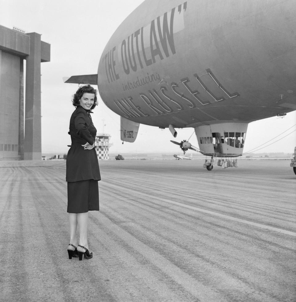 Actress Jane Russell stands before a surplus 167-foot Navy blimp that reads "Howard Hughes Daring Production The Outlaw" all over the sides in twenty-foot letters.