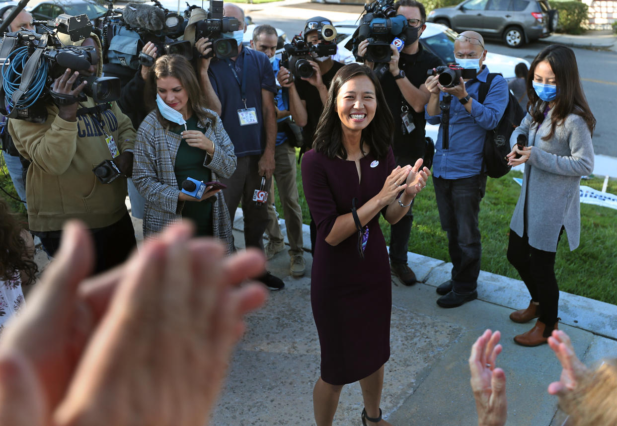 Michelle Wu stands in front of people with cameras, microphones and press badges while smiling and clapping with supporters, whose hands are visible in the foreground.