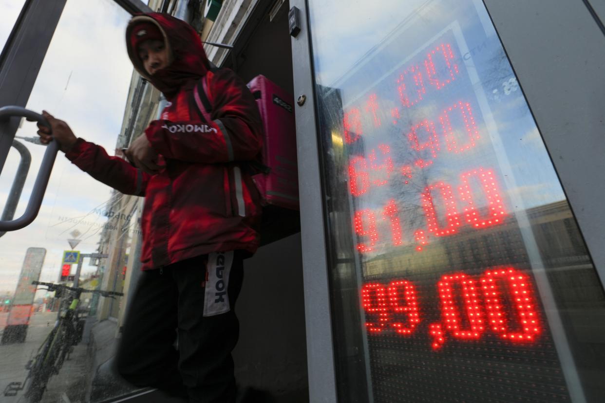 A food delivery man wearing a hooded jacket opens the door to leave an exchange office displaying currency rates.