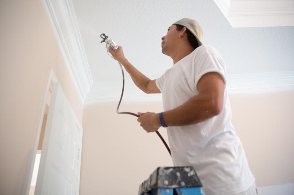 A man on a step ladder using a paint sprayer to paint the crown molding and ceiling of a room white.