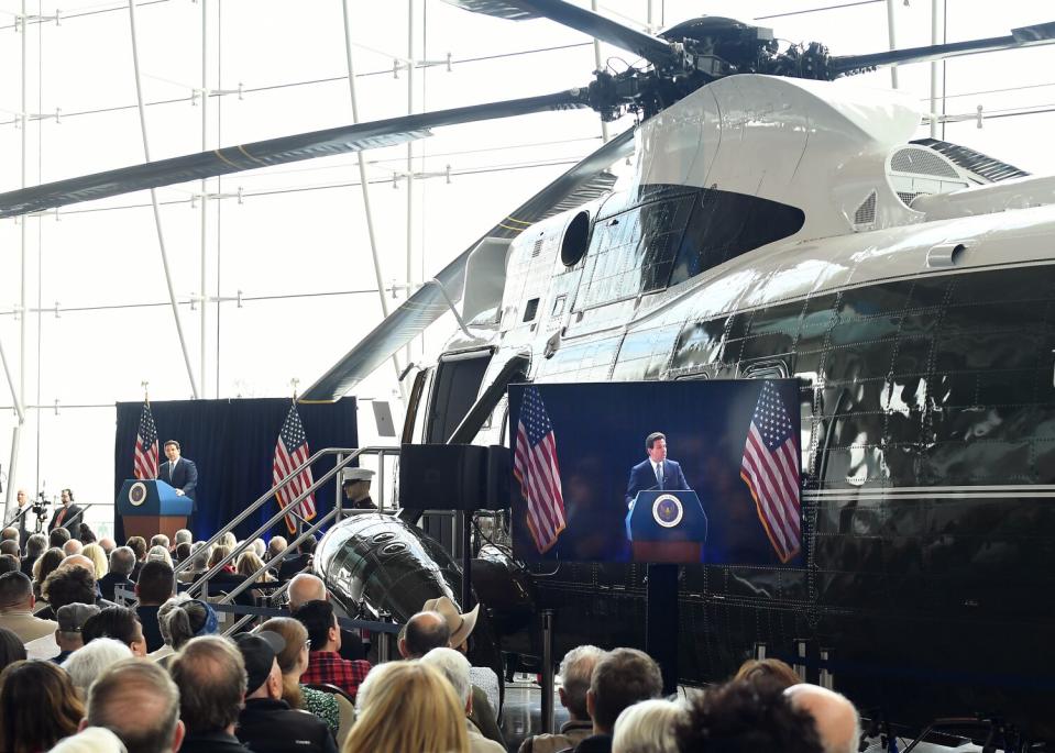 man in a suit and tie speaks from a podium standing in front of a retired presidential helicopter.