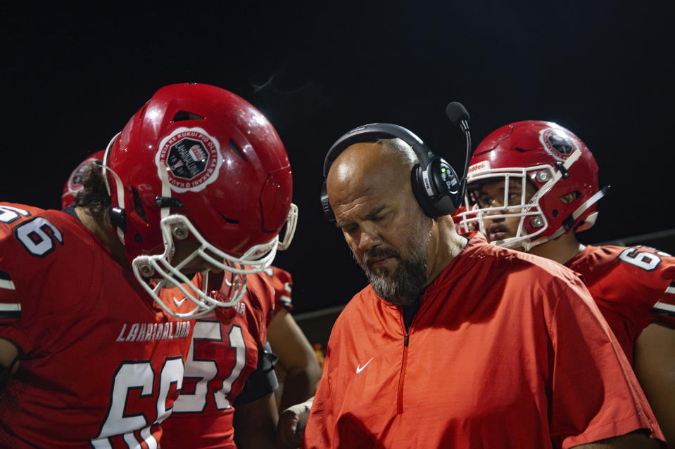 Lahainaluna High School football co-head coach Dean Rickard, front right, talks to players during a timeout during a game against Baldwin High School, Saturday, Oct. 21, 2023, in Lahaina, Hawaii. Lahainaluna’s varsity and junior varsity football teams are getting back to normal since the devastating wildfire in August. (AP Photo/Mengshin Lin)