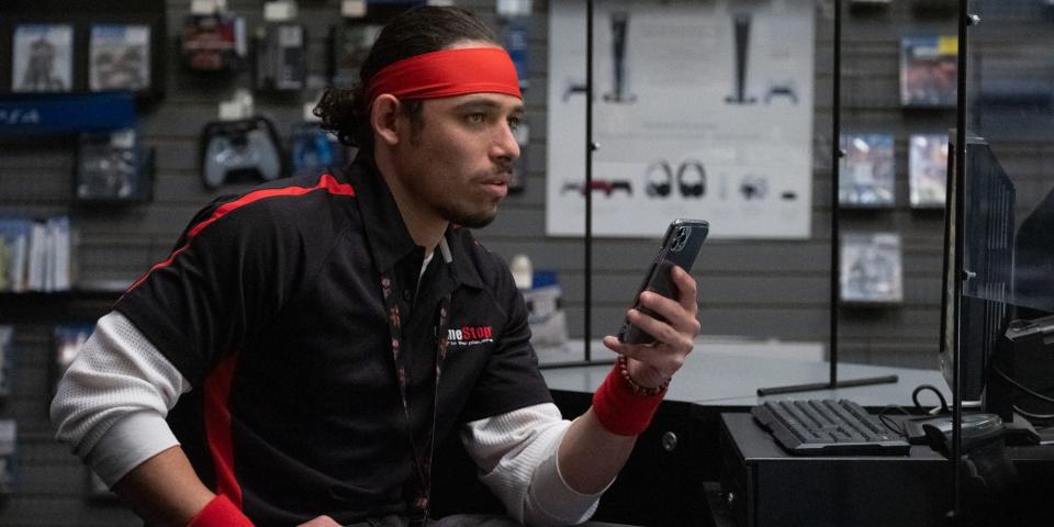 Anthony Ramos sits in a chair while on his phone as a GameStop employee in "Dumb Money."