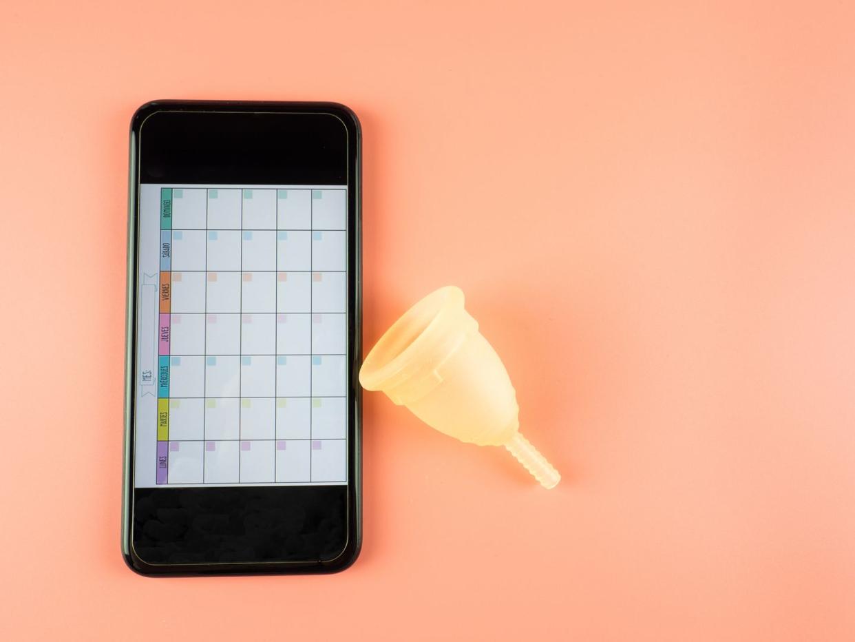 a menstrual cup next to a smartphone with a menstrual calendar mobile app on a pink background