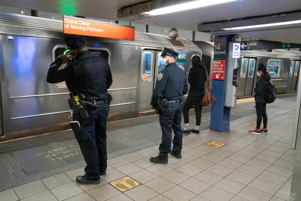 Two NYPD officers stand on a subway platform near two riders, all facing a train with closed doors.