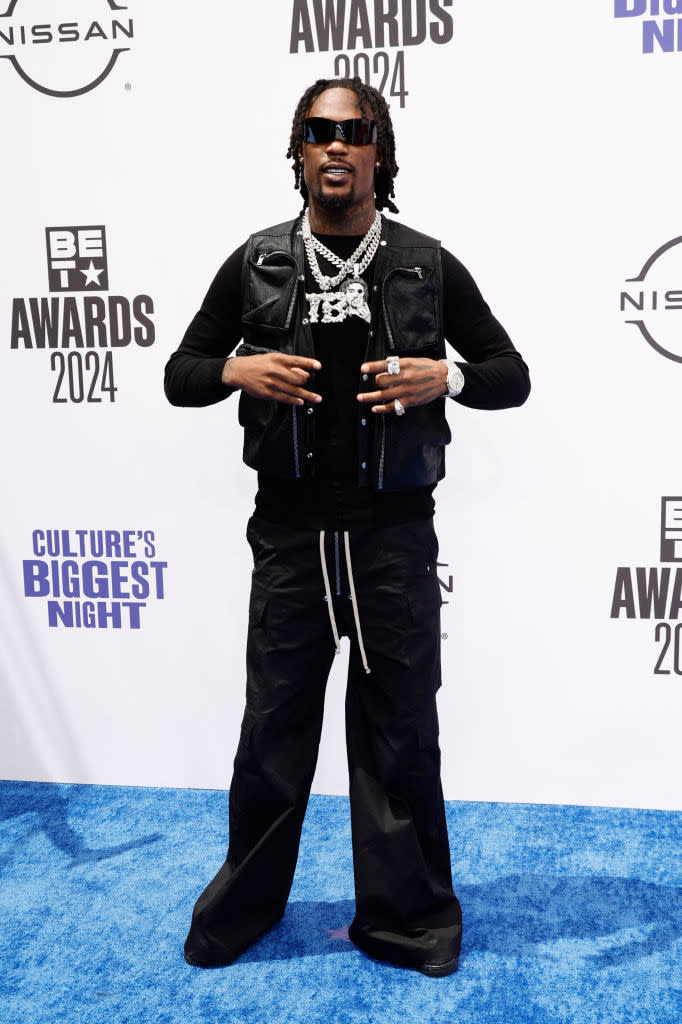 Hunxho poses on the red carpet at the 2024 BET Awards, wearing a black outfit with a sleeveless leather jacket, sunglasses, and multiple chains