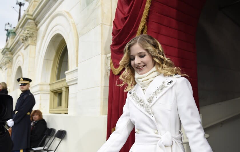 A woman in a dressy white winter coat looks down as she walks out of an archway