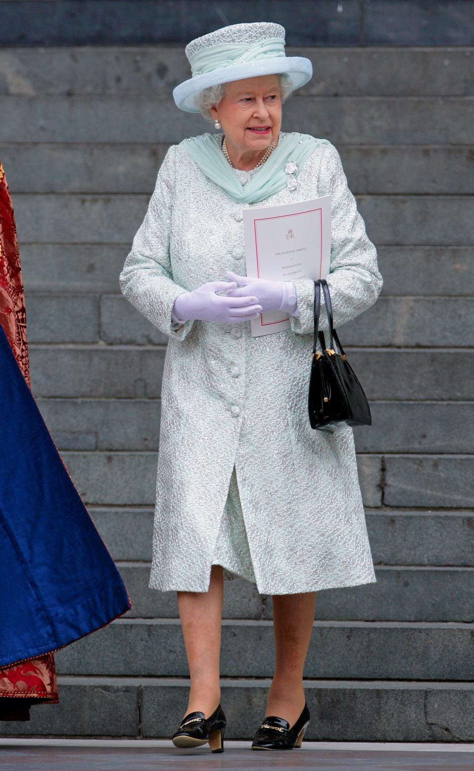 The queen walking in a pale blue and white textured overcoat with large buttons with a matching dress underneath. The collar of her coat is pale blue tulle with matches the ribbon around her wide-brimmed hat. She has on white gloves, a black purse and black low heels.