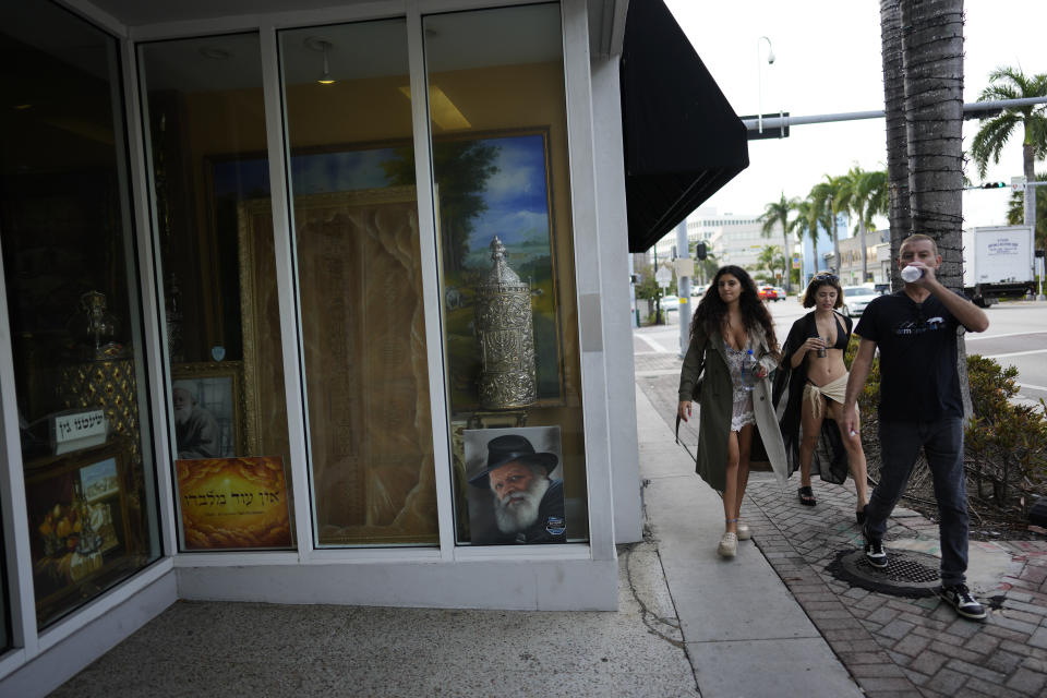 Women dressed for the beach walk past a business window displaying Jewish religious art, in Miami Beach, Fla., Friday, Dec. 1, 2023. The Jewish communities in and around Miami Beach for decades have been a welcoming home for Orthodox, Reform and secular Jews. But now, from Holocaust survivors to new moms, most here say daily life has been viscerally transformed by the Hamas attacks and the Middle East conflict that followed them. (AP Photo/Rebecca Blackwell)