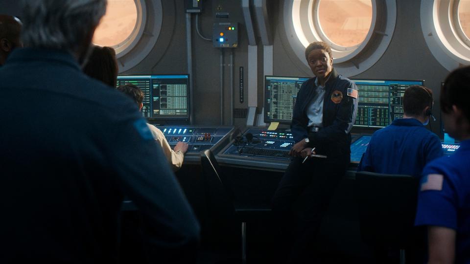 A woman with short black hair wearing a blue jumpsuit with an American flag patch on the shoulder is leaning against some space instrumental control panels in the control room. She has her hands crossed and is addressing a man in front of her (we can only see the back of his gray-haired head and blue jumpsuit). In the background you see more technical screens and control monitors manned by staff.