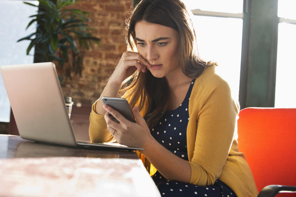 Caucasian woman sat at desk with laptop open, looking worriedly at mobile phone