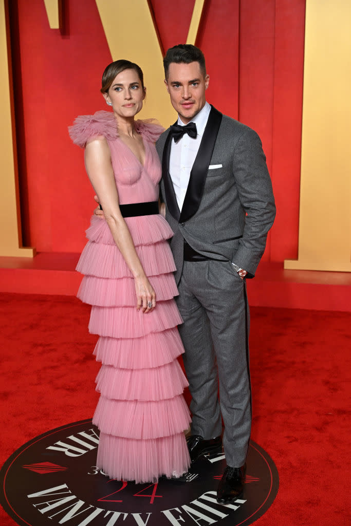 Two people posing together at the Vanity Fair event, one in a layered ruffled dress and the other in a gray suit with a bowtie