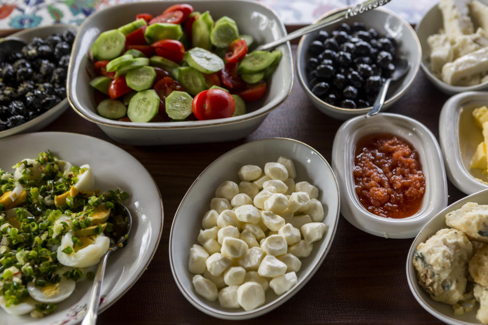 Several bowls of fresh foods including cucumber and tomato salad, black olives, cheese balls, chopped egg salad, tomato sauce, butter, and bread rolls