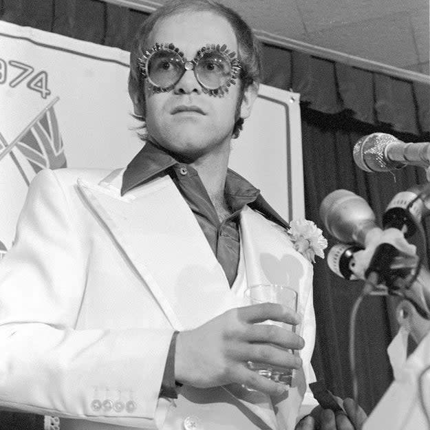 Elton John, in an ornate suit and unique glasses, stands at a press conference holding a drink with microphones in front