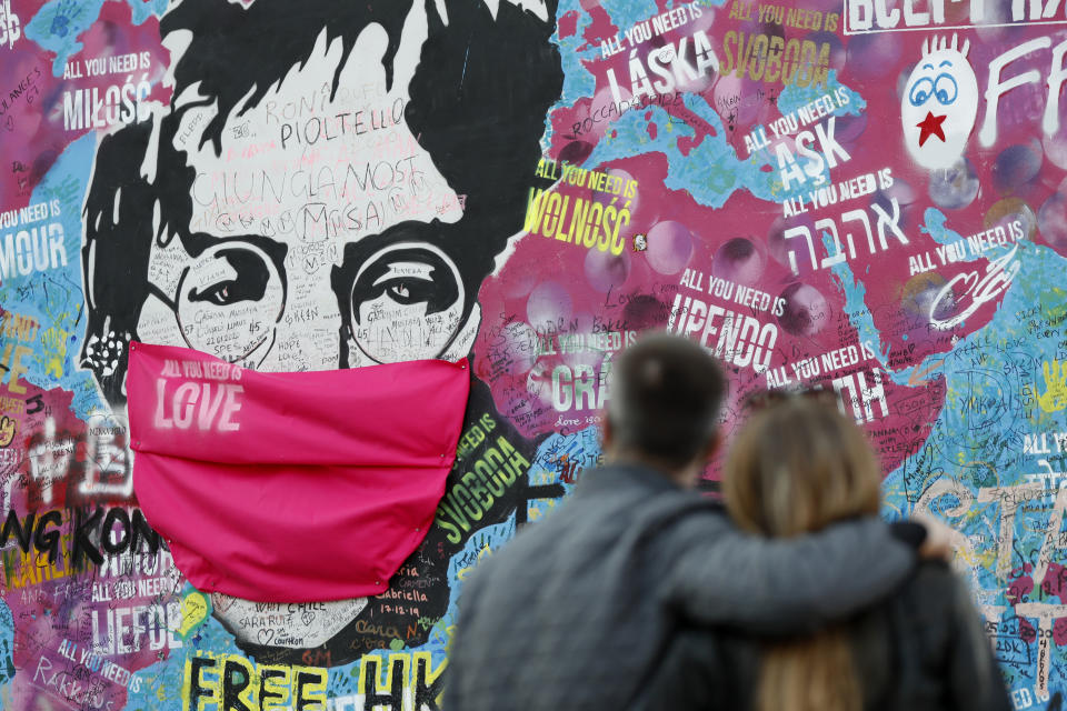 FILE - A couple look at the "Lennon Wall" with a face mask attached to the image of John Lennon, in Prague, Czech Republic, on April 6, 2020. Like so many other events in the year of coronavirus, an annual tribute to John Lennon held in its adopted city of New York will go online. The five-hour event will be streamed for free on Lennon's birthday, October 9, starting at 7 p.m. Eastern time on the LennonTribute.org website. It will feature recorded performances from Patti Smith, Rosanne Cash, Natalie Merchant, Jackson Browne, Jorma Kaukonen and others. (AP Photo/Petr David Josek)