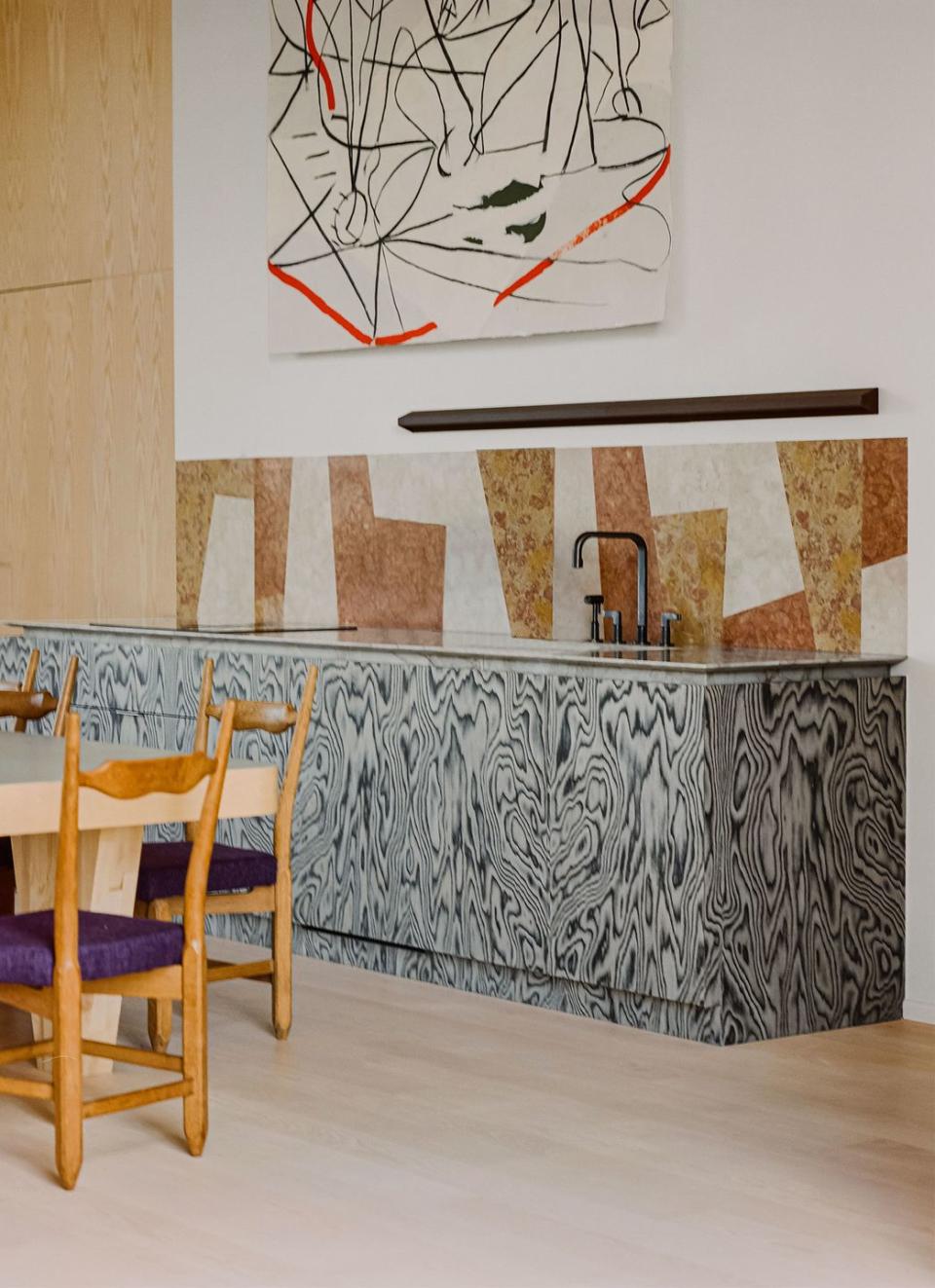 a kitchen has a heavy wood table and wood chairs with dark purple seat cushions, a sink and counter with swirly gray and white cabinets, a backsplash of irregularly shaped tiles, and a large abstract artwork