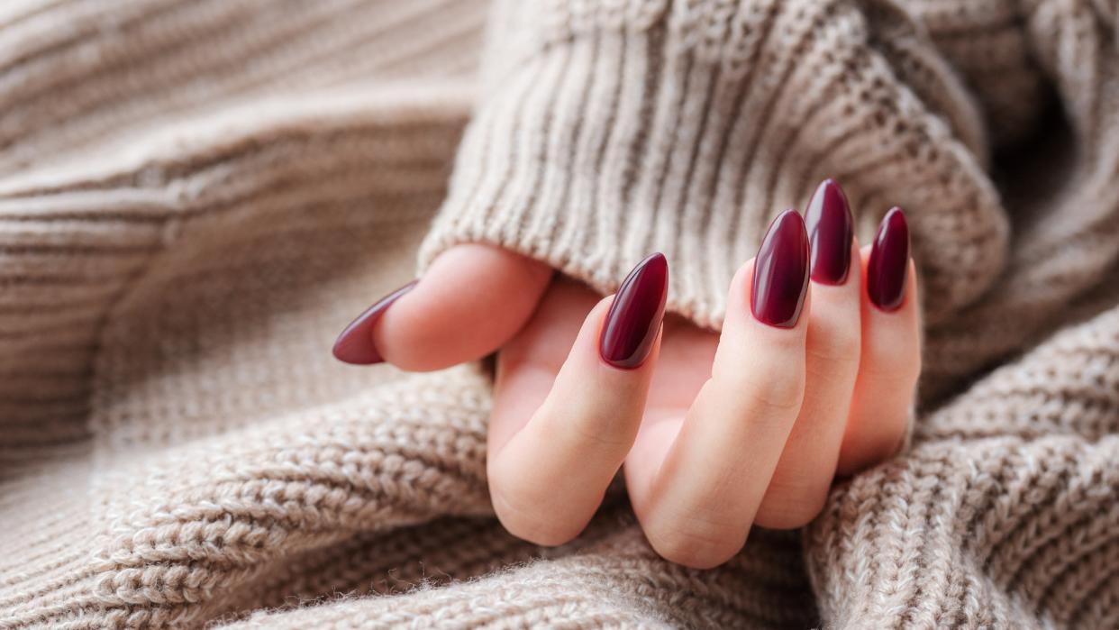  A hand with long, oval shaped dark red nails on a cream, knitted background. 