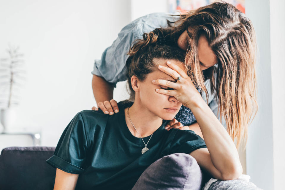 Two people: one is sitting with a hand covering their face, looking sad, while the other is standing behind, offering comfort with a hand on the shoulder.