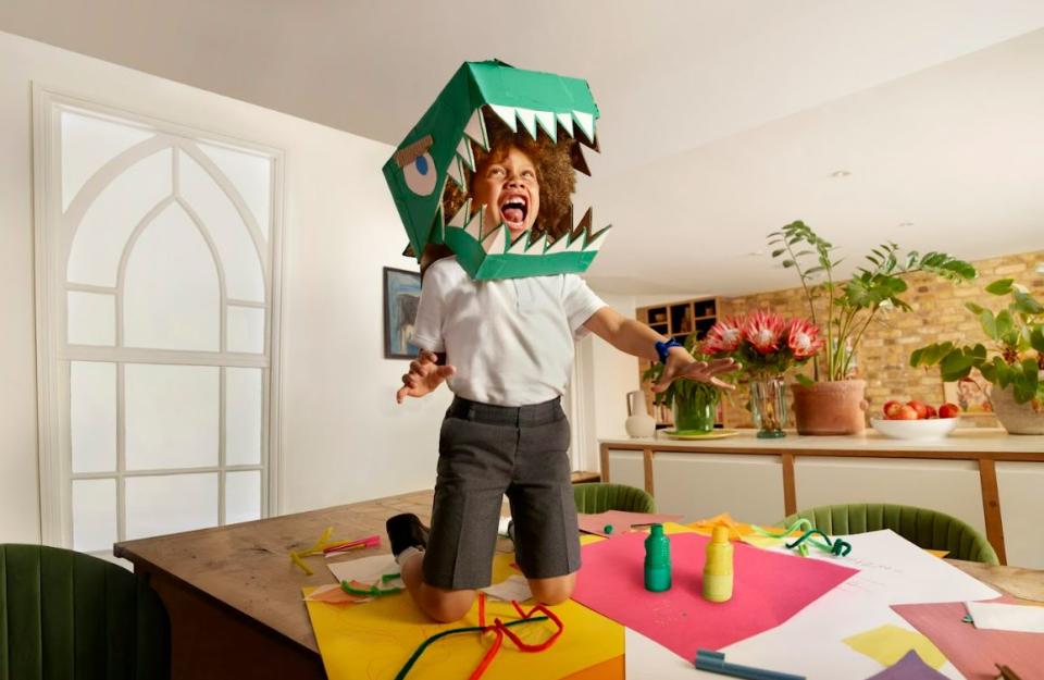 A smiling child in school uniform kneels on a school table, wearing a paper mache crocodile mask