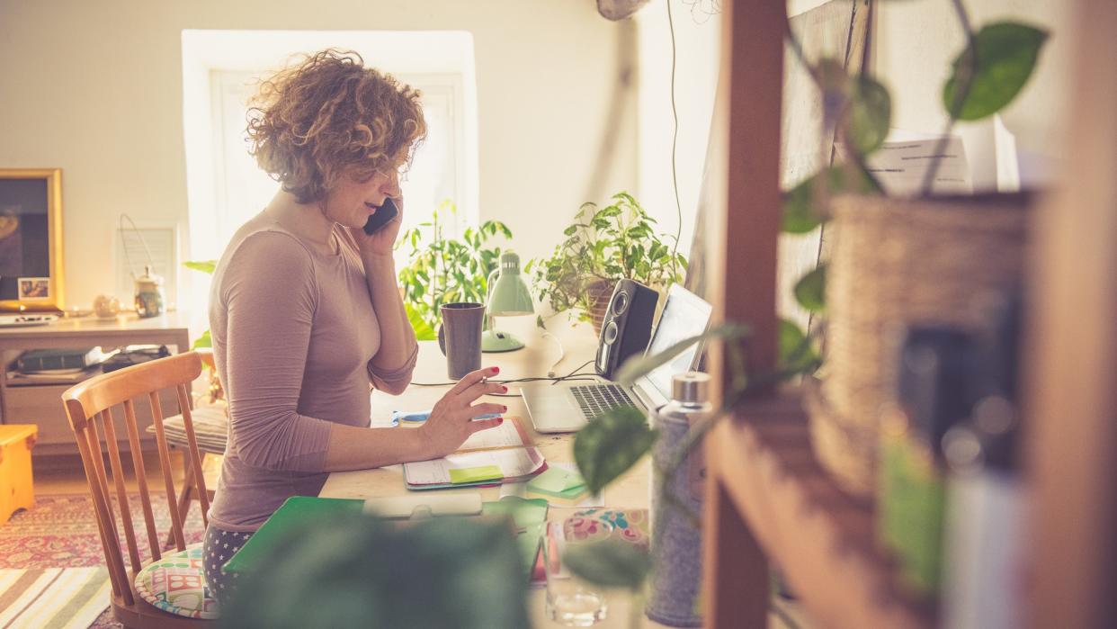 Woman working from home, using phone, sitting on the chair and writing into the book.