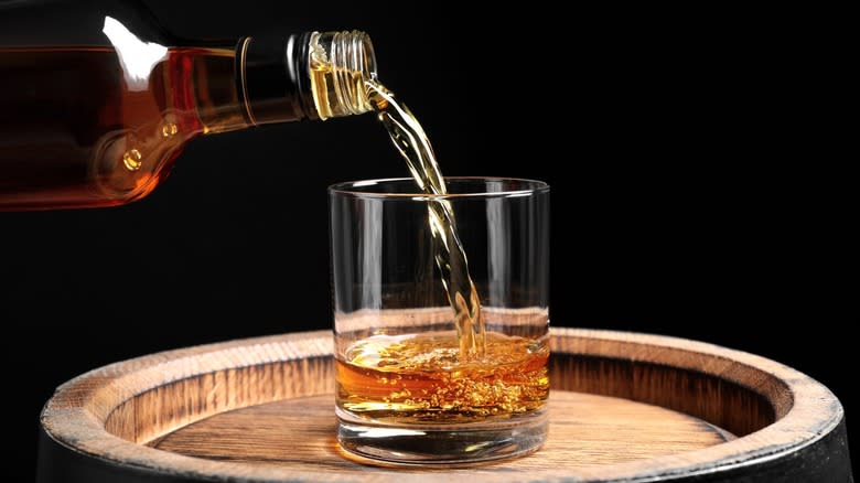 Whiskey being poured from a bottle into a glass on a barrel